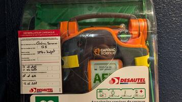 A defibrillator in a plastic case with a maintenance label and emergency instructions.