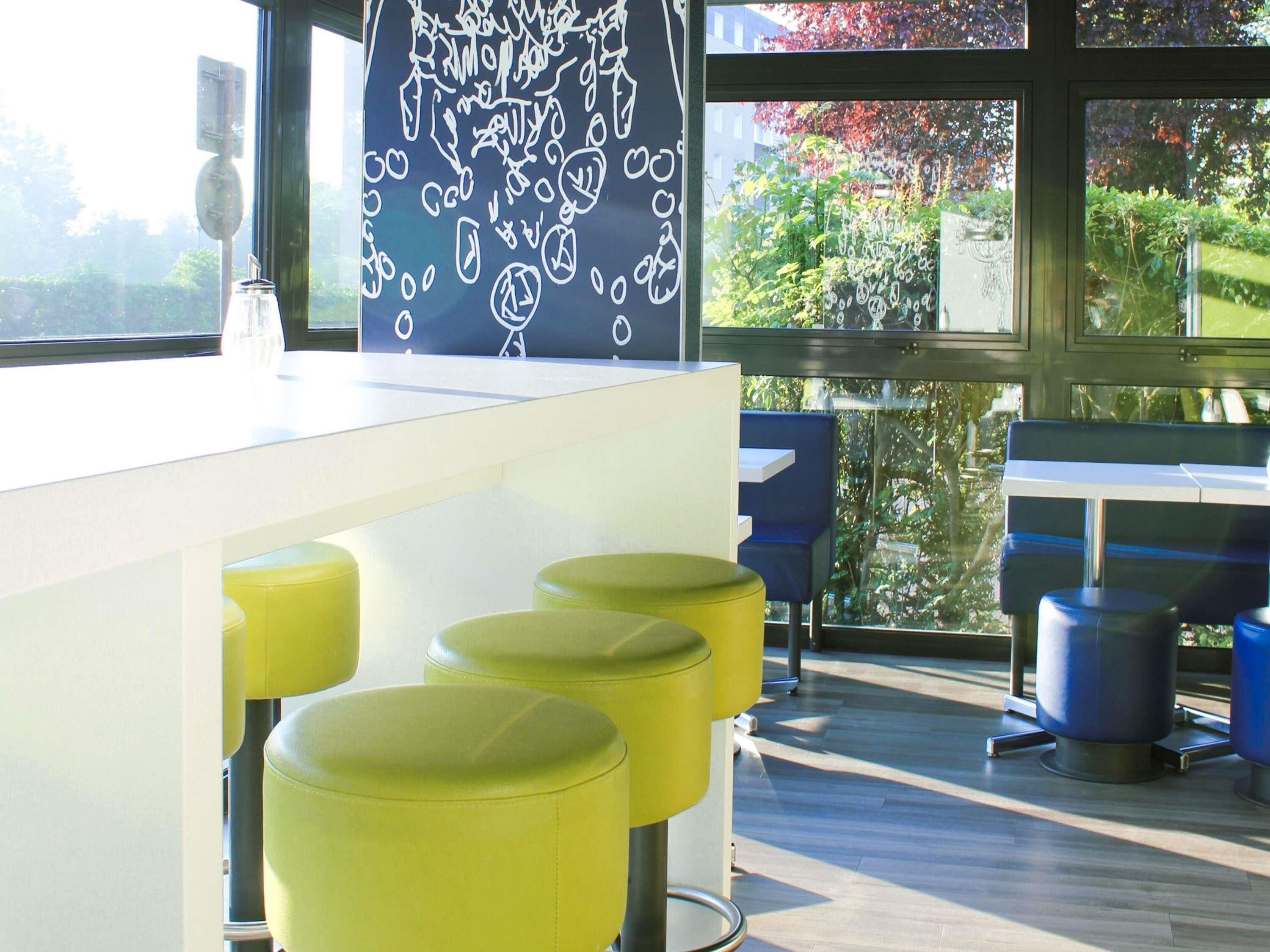 Un espace bar moderne avec un comptoir blanc, des tabourets jaunes et une grande fenêtre donnant sur un paysage verdoyant.