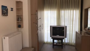 A living room with a television on a stand, a radiator, a shelf with decorative items, and a window with curtains. - Luxury Maisonette in Halkidiki (Kassandra)
