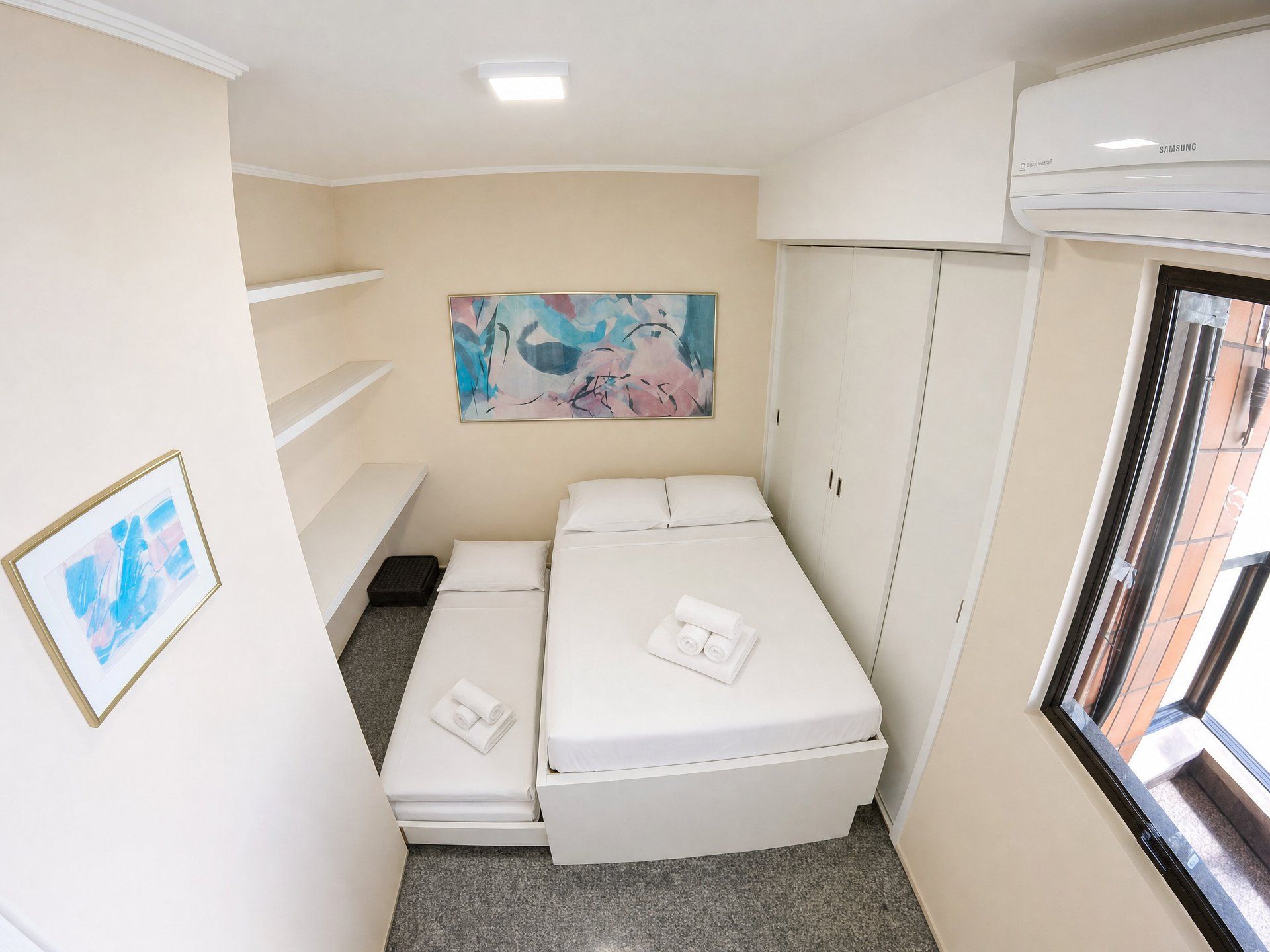Quarto de hotel pequeno com cama, guarda-roupa, um quadro na parede e uma janela.