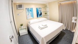 Quarto de hotel com cama, mesa de cabeceira, ar condicionado, arte abstrata na parede, prateleira de vidro com plantas e janela com cortinas.