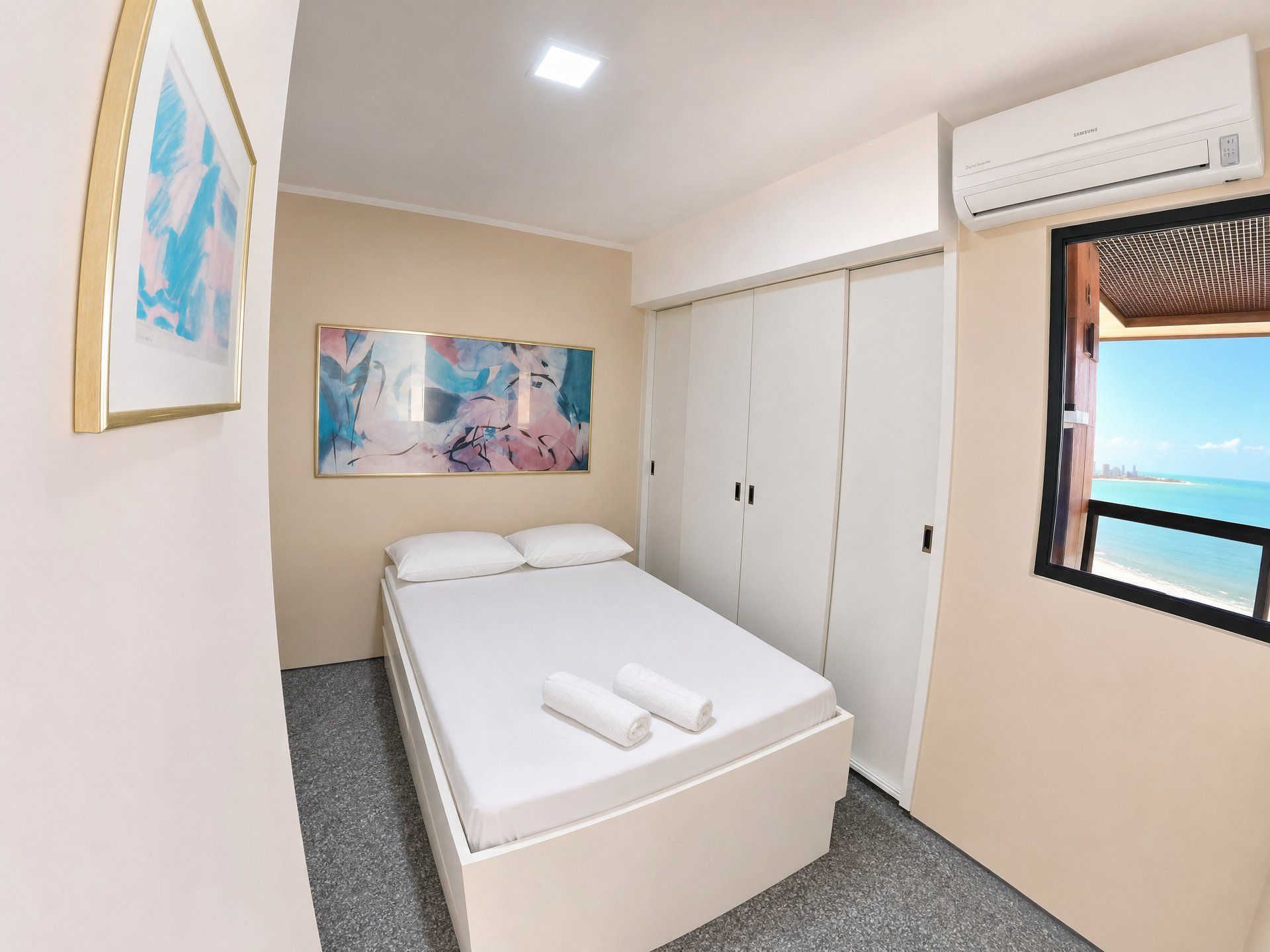 A hotel room with a bed, a painting, a wardrobe, an air conditioning unit, and a window with a view.