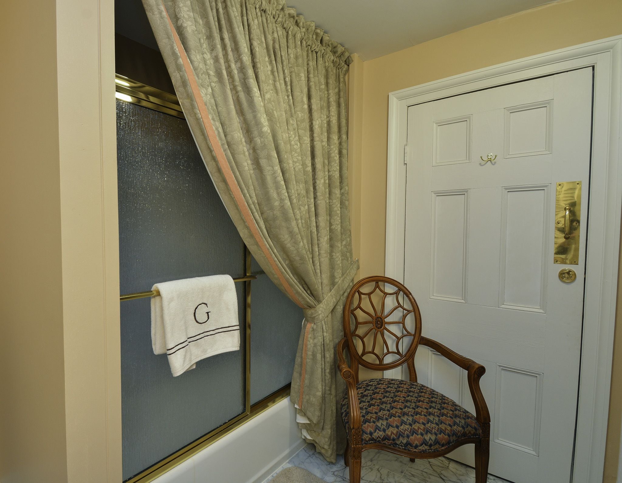 A bathroom with a towel rack holding a towel with a letter 'G', a wooden chair, and a white door with a brass handle.