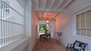 A covered outdoor corridor with a brick floor, white walls, a white railing, a table with chairs, a bench, and a hanging light fixture.