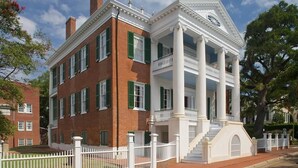 A two-story brick building with white columns and a balcony, surrounded by a white fence.