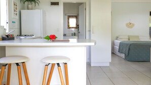 A modern kitchen with white countertops, wooden stools, a clock, and a potted plant. - Makayla Palms (Rarotonga)