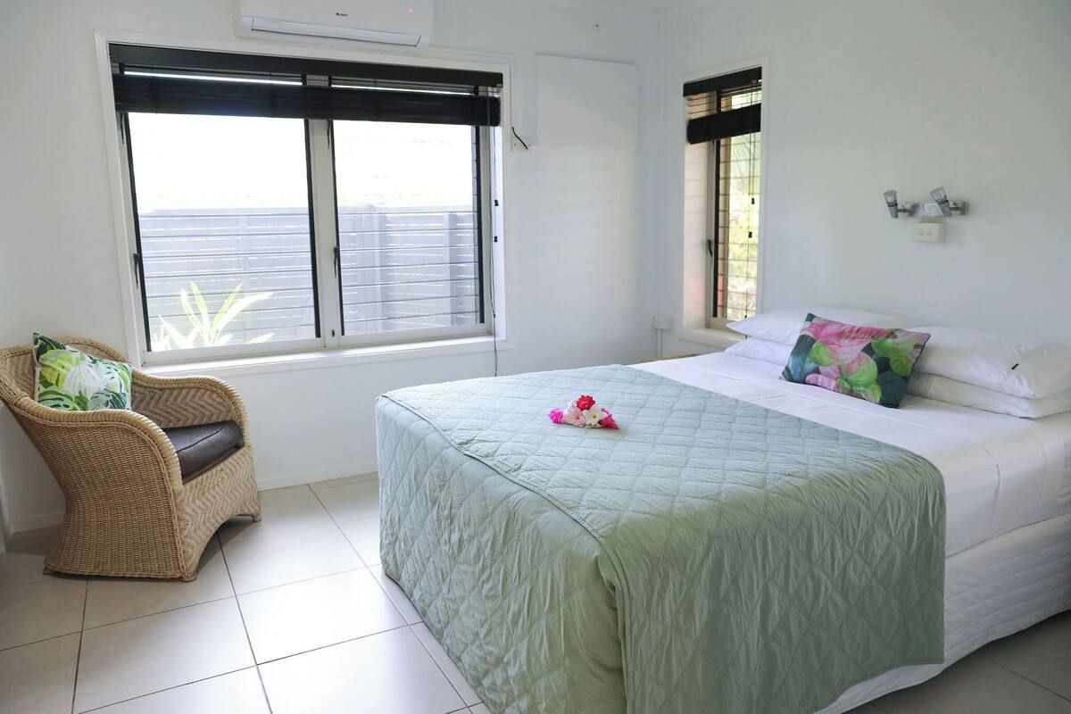 A bedroom with a bed, a wicker chair, a large window, and a view of the outside.