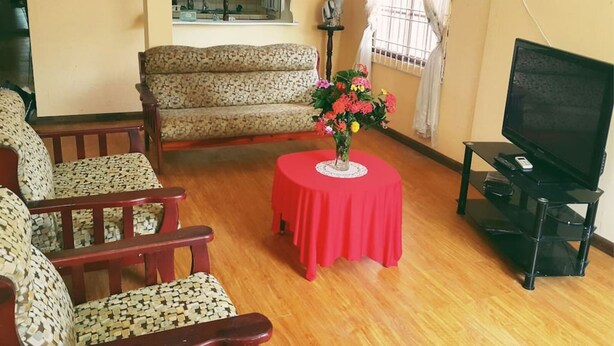 Un salón con un sofá, dos sillones, una mesita roja con un jarrón, un televisor sobre un mueble bajo y una ventana con cortinas.