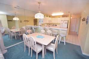 A dining area with a table and chairs, a kitchen in the background, and a door leading to another room.