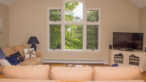A living room with a beige sofa, a coffee table, a TV on a white cabinet, and a large window with a view of trees. - Sun, Sand, and Serenity: Only 3 peak weeks remaining - Includes Private Beach (Eastham)