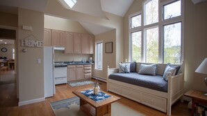 A spacious living room with a large window, a cozy seating area, and a kitchen in the background.