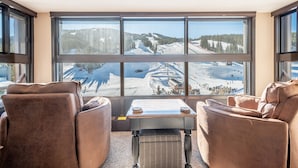 A room with a large window offering a view of a snowy landscape and a ski slope. There are two brown leather chairs and a small table with a book and a cup. - The best view in Center Village.  Perfect for Concerts and Skier watching!!! (Copper Mountain)