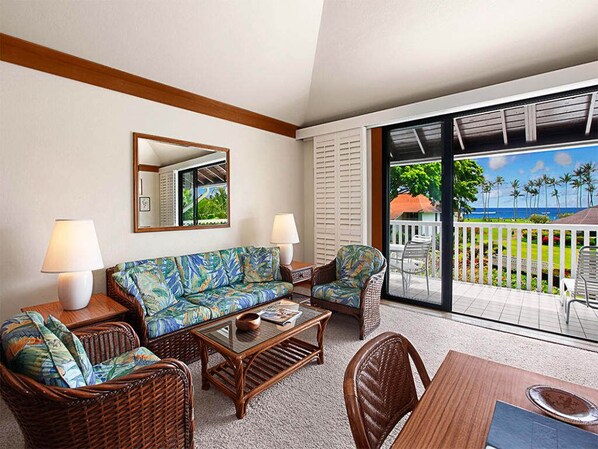 A living room with a sofa, chairs, and a coffee table. There is a balcony with a view of palm trees and the ocean. - Poipu Perfection w/WiFi, Kitchen, Lanai, Ceiling Fans, Flat Screen+DVD–Kiahuna 2073 (KOLOA)