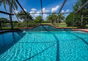 A clear blue pool with a tiled edge, surrounded by a metal fence and palm trees. - Airy house with heated waterfall pool overseeing golf course, minutes from beach (Port St. Lucie)