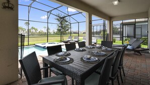 A covered patio with a dining table set for six, overlooking a swimming pool and a grassy area.