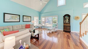 A spacious living room with a high ceiling, hardwood flooring, a large window, and a ceiling fan.