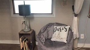 Ein Zimmer mit grauem Sitzsack, einem kleinen Beistelltisch mit Lampe und einem Fenster mit Vorhängen.