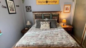 Ein Bett mit Paris-Motiv auf der Bettdecke, ein Nachttisch mit Lampe und ein Wandschild mit der Aufschrift "Always Kiss Me Goodnight".