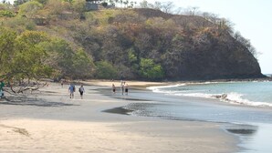 A beach with people walking along the shore, a rocky cliff with sparse vegetation, and a clear sky.