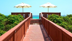 Eine Holzbrücke führt zu Strandstühlen, umgeben von viel Grün.