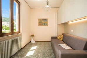 A room with a radiator, a sofa with a cushion, a small table with a glass top, a painting on the wall, and a chandelier. - Villa Alberti Appartamento Emma, Salò, Italy (Salò)
