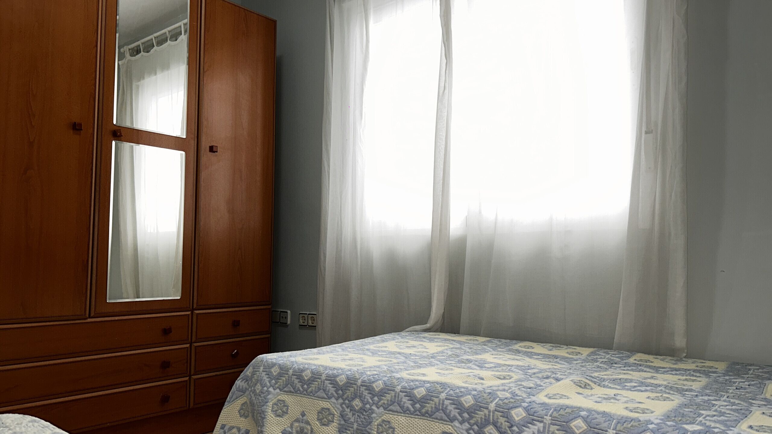 Un dormitorio con un armario de madera, una cama con ropa de cama estampada y una ventana con cortinas de gasa.