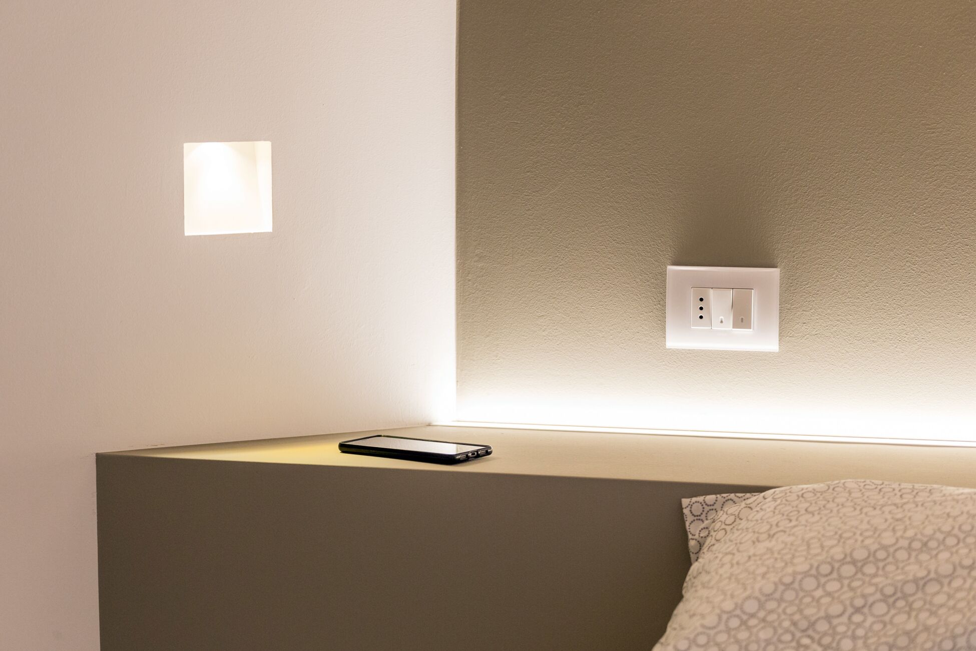 A bedroom with a bedside table, a smartphone, a wall socket, and a light switch.