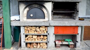 Ein gemauerter Außenofen mit Feuerraum, Holzablage und einem kleinen Tisch mit Besen und Sonnenschirm.
