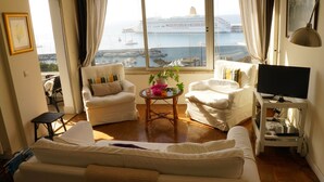 Un salon comprenant un canapé, deux fauteuils, une petite table ornée de fleurs, un téléviseur posé sur un meuble, et une vue sur un navire de croisière à travers de grandes fenêtres.