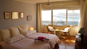 Une chambre d’hôtel avec un lit, deux fauteuils en osier, une petite table et une vue sur un navire de croisière et un port de plaisance.