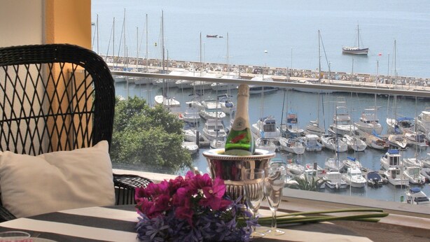 Une table dressée avec une bouteille, des fleurs et des assiettes, donnant sur une marina où sont amarrés des bateaux.