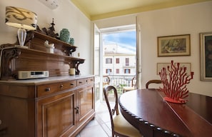A dining area with a wooden table and chairs, a cabinet with decorative items, and a view of buildings through an open door. - In Alghero in the historic center on the square, large and bright apartment (Alghero)