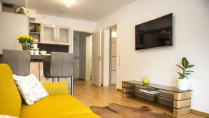Uma sala de estar moderna com um sofá amarelo, uma mesa de centro de vidro e uma TV na parede.