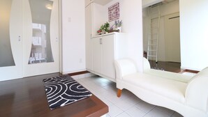 Un salón moderno con un sofá blanco, una mesa de centro de madera y una alfombra decorativa. Se ve un armario con plantas y una escalera al fondo.