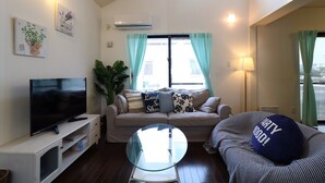 Una sala de estar con un televisor de pantalla plana, una mesa de centro de vidrio, un sofá con cojines estampados y un ventanal con cortinas.