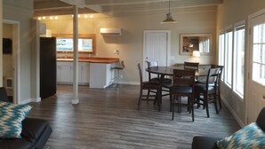 Un amplio espacio de sala con una mesa y sillas de comedor, una cocina al fondo y ventanas grandes que dejan entrar luz natural.