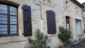 Een stenen gebouw met twee ramen, waarvan er één luiken heeft en de ander een klein balkon. Voor het gebouw staan bloemen en planten in potten.