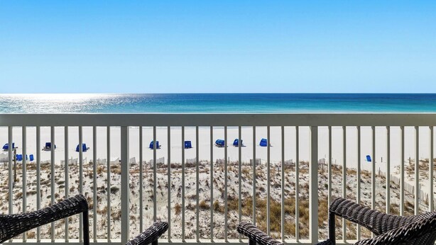 Un balcone con sedie in vimini che si affaccia su una spiaggia con sedie blu e una recinzione.