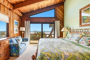 Ein Schlafzimmer mit einem Bett, einer hölzernen Kommode, einem Sofa und einem großen Fenster mit Blick auf den Strand.