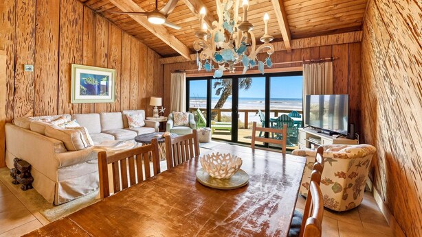 Interior de madera con una mesa de comedor, sillas, un sofá, una araña y vista a la playa.