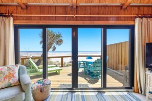 Ein Zimmer mit einer großen Schiebeglasdoor, die auf eine Terrasse mit Gartenmöbeln und Blick auf den Strand führt.