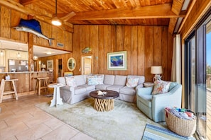 A wooden interior with a sofa, a round coffee table, a kitchen area, and a large window.