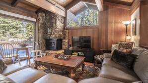 Un salon chaleureux avec une cheminée en pierre, un plafond en bois et un grand téléviseur. On y trouve des coins salon confortables et une vue sur l’extérieur.