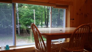 A wooden dining table with four chairs, large windows with a view of a forest, and a wooden wall.