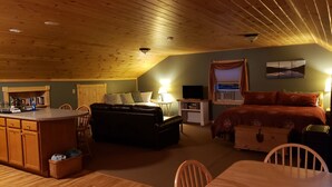 A cozy room with a wooden ceiling, a bed with a red blanket, a small table with chairs, a sofa, a TV, and a window with curtains.