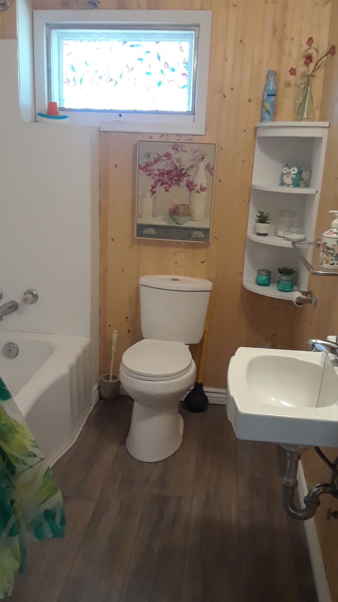 A bathroom with a toilet, sink, and bathtub. There is a window above the toilet, a shelf with decorative items, and a wooden floor.