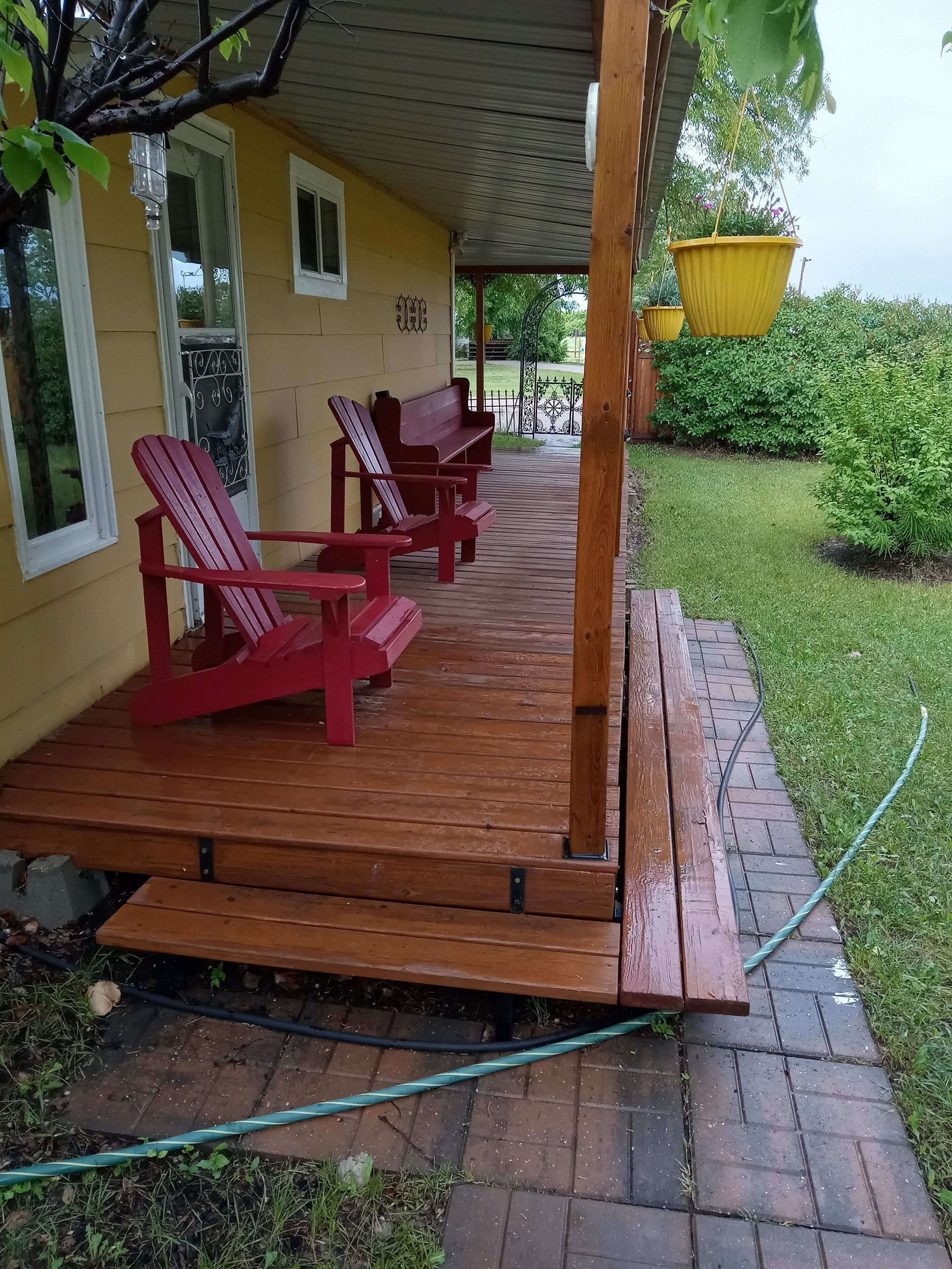 A covered porch with red Adirondack chairs, a yellow house, and a green hose on the ground.