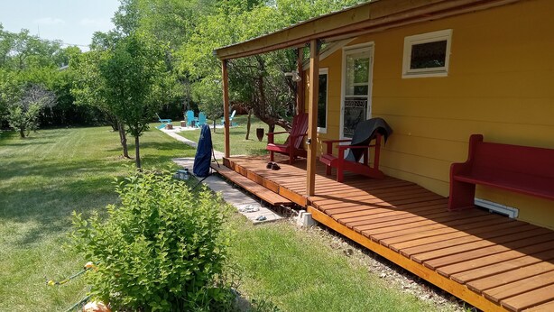 Una terraza de madera con mobiliario exterior, un porche cubierto y un pequeño jardín con una maceta y rocas.