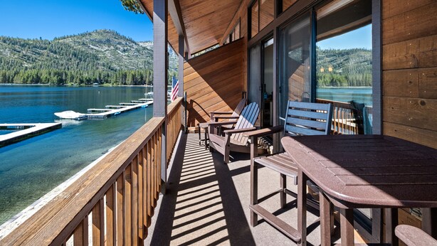 A wooden deck with a table and chairs overlooking a lake with boats, a mountain in the background, and an American flag. - Donner Vista: Lakefront Condo with Boat Slip, Sandy Beach and Unbeatable Views (Truckee)