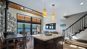 A spacious kitchen with a large island, wooden flooring, and a stone wall. There is a stone fireplace, a dining area with chairs, and a view of the outdoors through large windows. - Donner Vista: Lakefront Condo with Boat Slip, Sandy Beach and Unbeatable Views (Truckee)